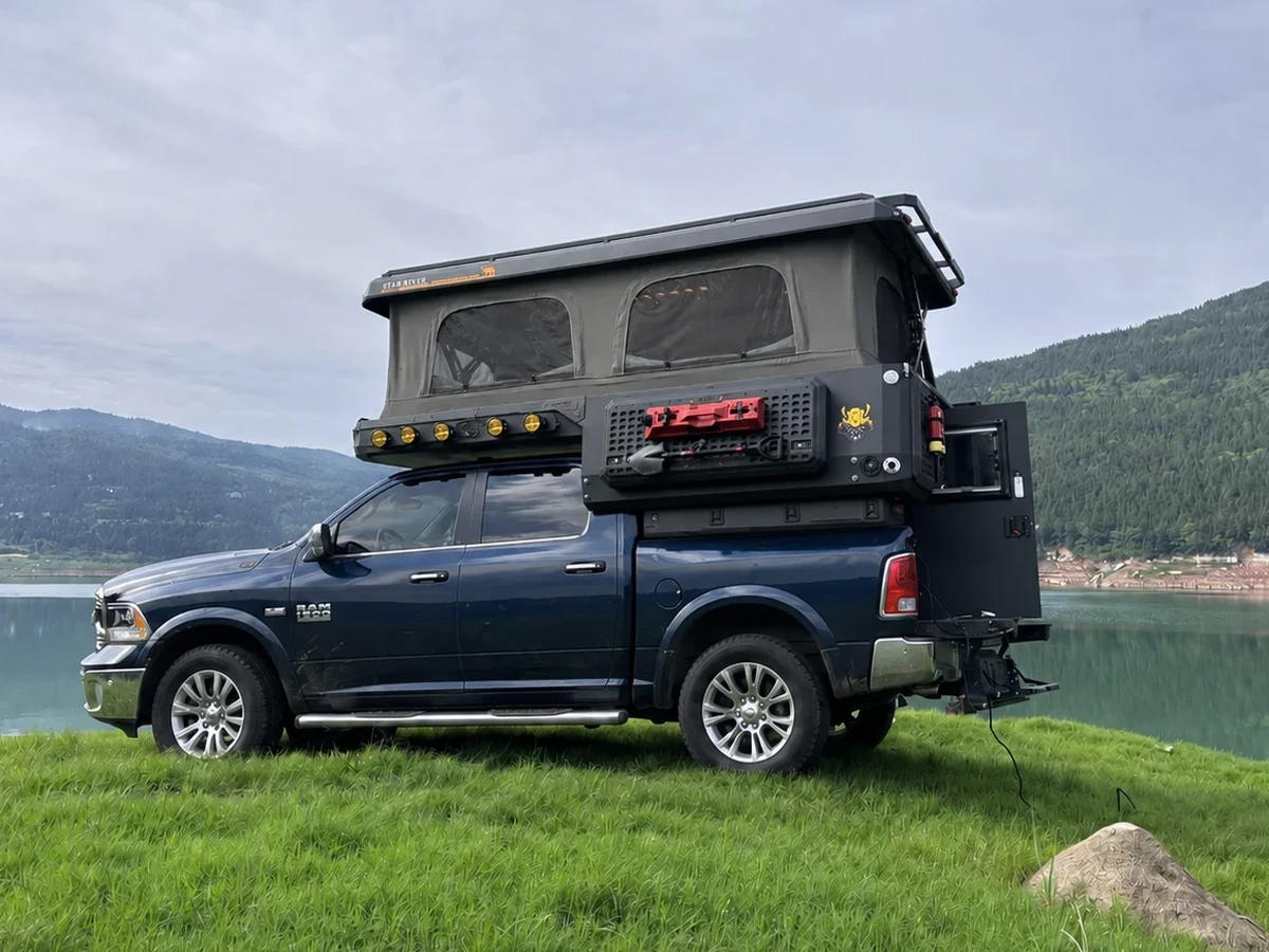 Blue RAM with a SUNHOMEE slide-in camper, roof raised beside a mountain lake